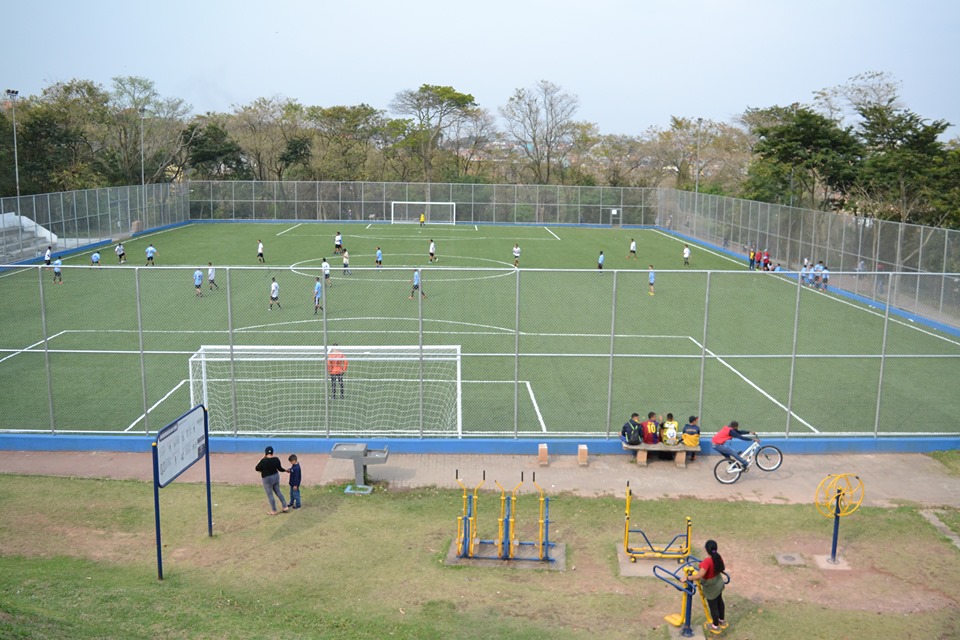 Foto feita com drone, mostra o campo de futebol completamente repaginado (grama verdinha, linhas bem marcadas, traves novas pintadas de branco e grades de aço). Dentro do campo, dois times de futebol masculinos se enfrentam. Um de azul e outro de branco.